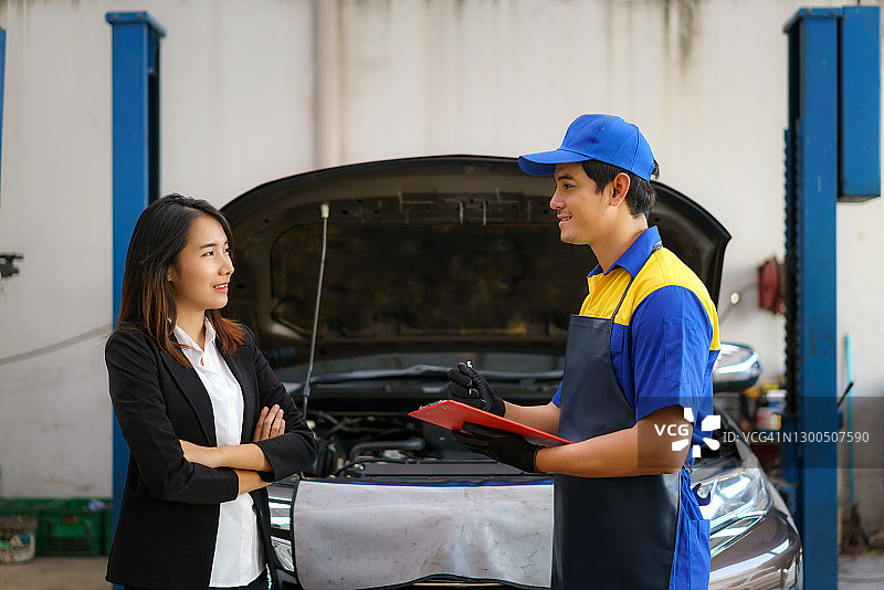 身穿蓝色制服的亚洲汽车修理工正在检查现代汽车，并向顾客解释维修事宜，汽车维修服务概念图片素材