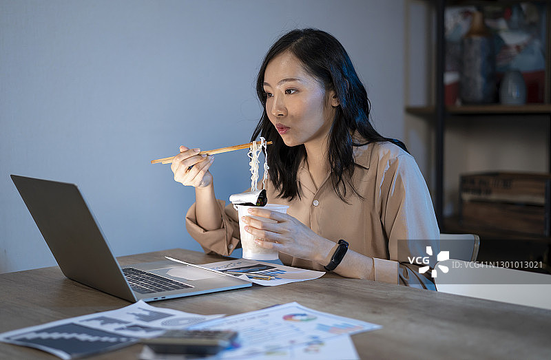 亚洲年轻女人夜晚在办公室加班吃泡面用电脑图片素材