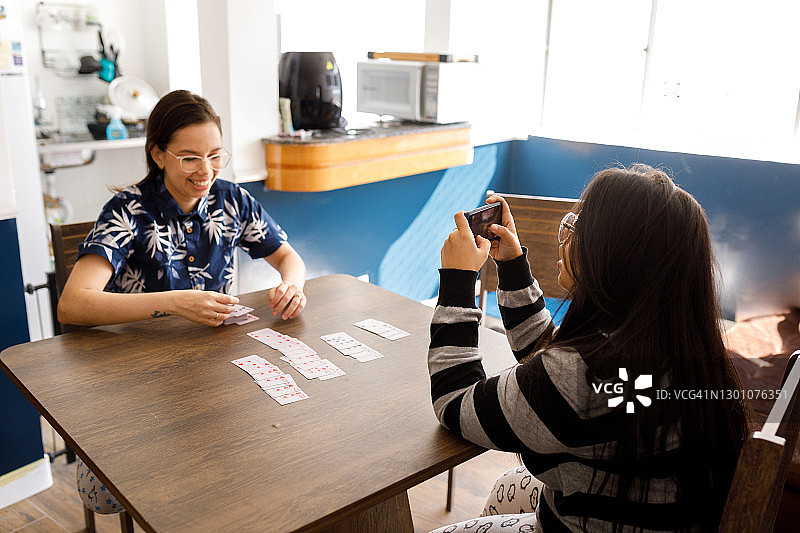 坐在桌旁，用智能手机和伴侣玩纸牌游戏的微笑年轻女人们图片素材