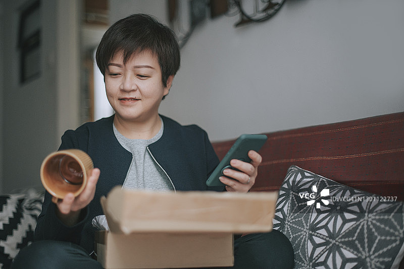 中国女人坐在沙发上拆开网购的咖啡杯和盘子，看着手机图片素材
