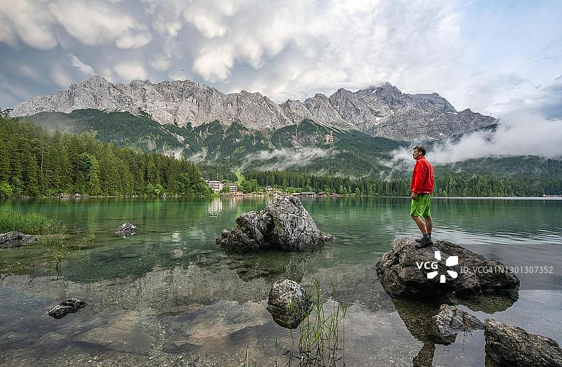 年轻男子站在岸边岩石上，楚格峰山脉前的艾布湖，德国巴伐利亚上巴伐利亚格赖瑙附近图片素材