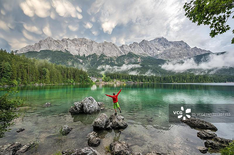 年轻男子在艾布湖岸边的岩石上张开双臂，背景是楚格峰山脉图片素材