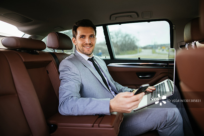 坐在汽车后座上用笔记本电脑和智能手机的英俊商人图片素材