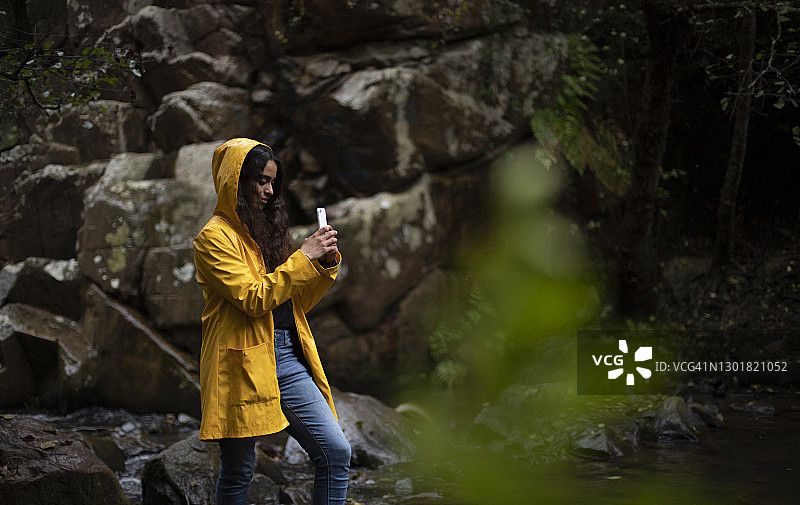 穿着黄色雨衣的女人在森林里用智能手机拍照图片素材