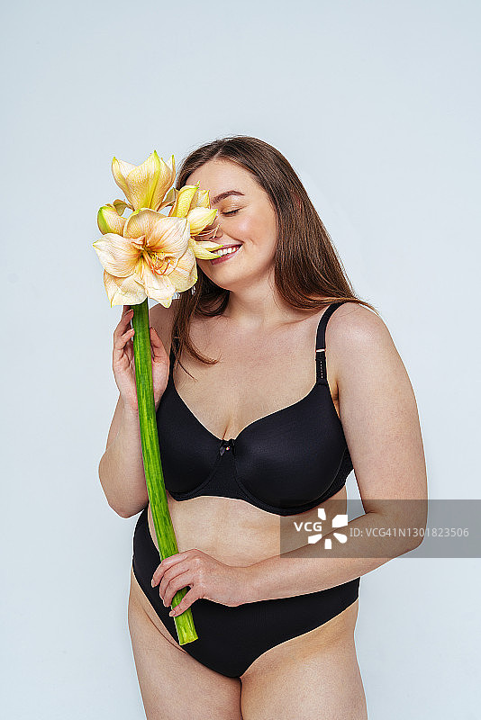 微笑的女模特穿着黑色比基尼，在白色背景下闻花图片素材