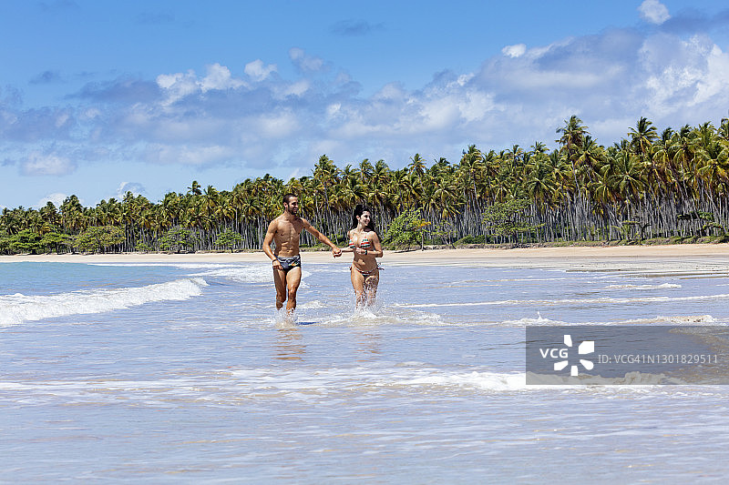 一对穿着泳装的迷人年轻夫妇在热带海滩的波浪中奔跑图片素材