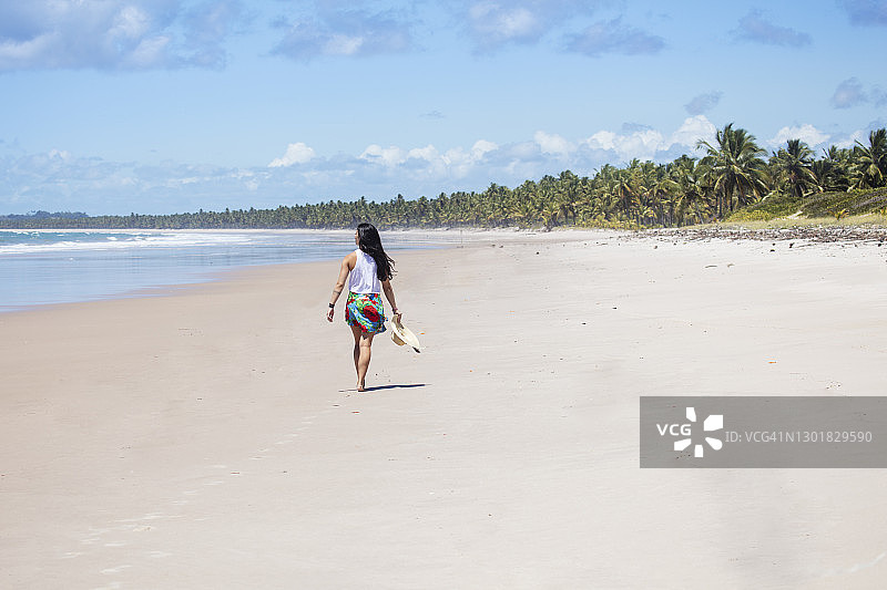 穿着海滩服装，拿着帽子走在热带海滩上的年轻女子图片素材
