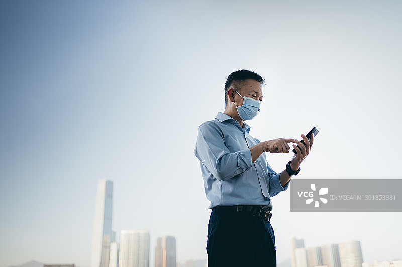戴防护口罩，在香港海港使用智能手机的亚洲年长商人图片素材