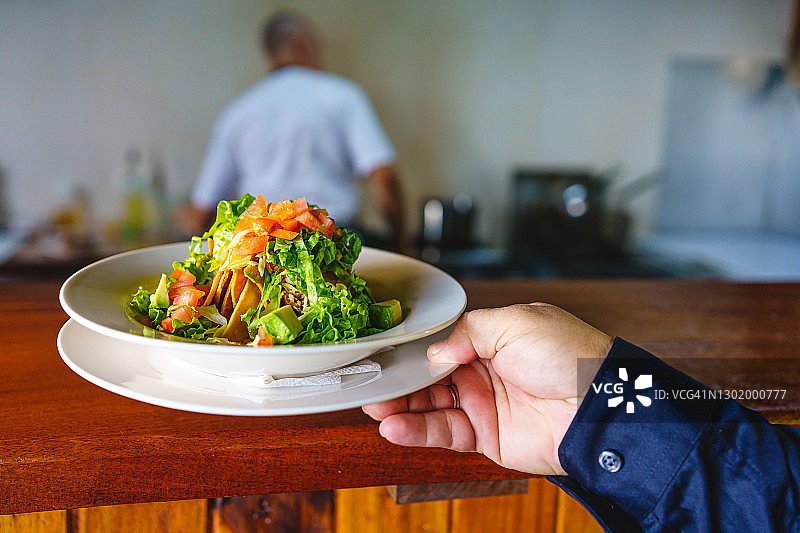 在餐厅里，服务员的手端着一盘沙拉，背景是厨师图片素材