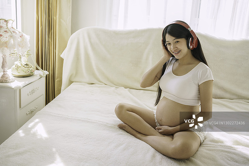 身穿白色衣服的亚洲孕妇幸福地坐在床上图片素材