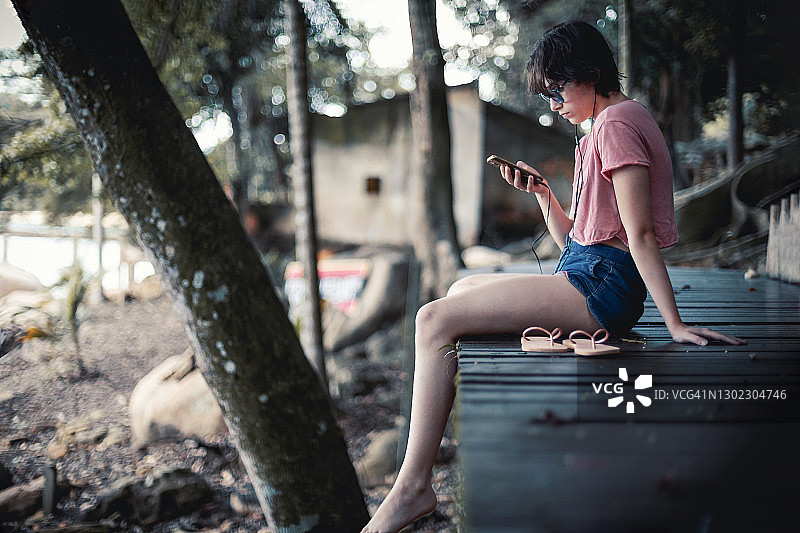 小女孩坐在大自然的木制码头上，用智能手机听音乐图片素材