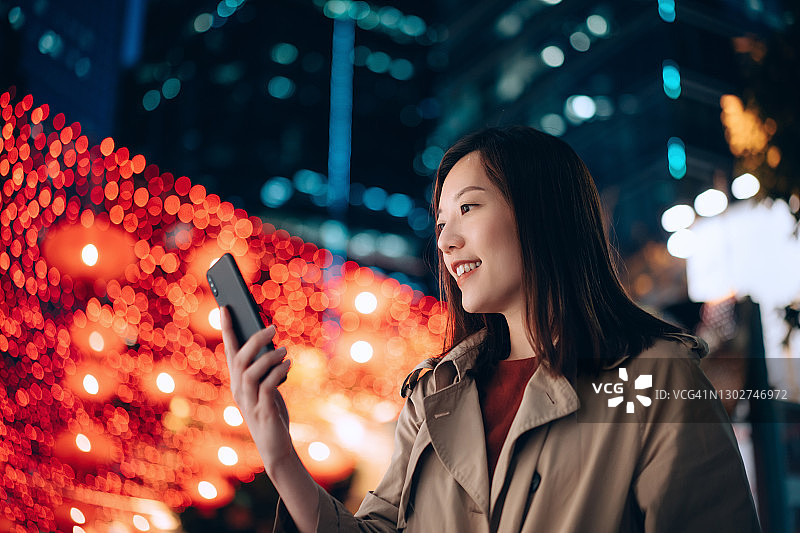 年轻亚洲女商人在夜晚城市中使用智能手机的仰视视角肖像，背景是灯火通明的城市商业建筑和充满活力的城市街灯。移动办公图片素材