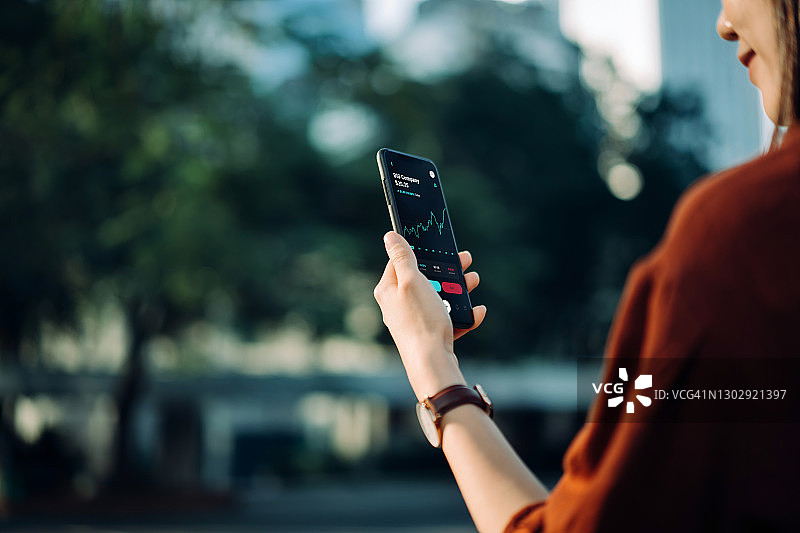 在中央商务区的办公园区，一位亚洲年轻女商人使用手机App查看金融股市分析，沐浴在阳光下，周围是绿色的自然植物。移动办公图片素材