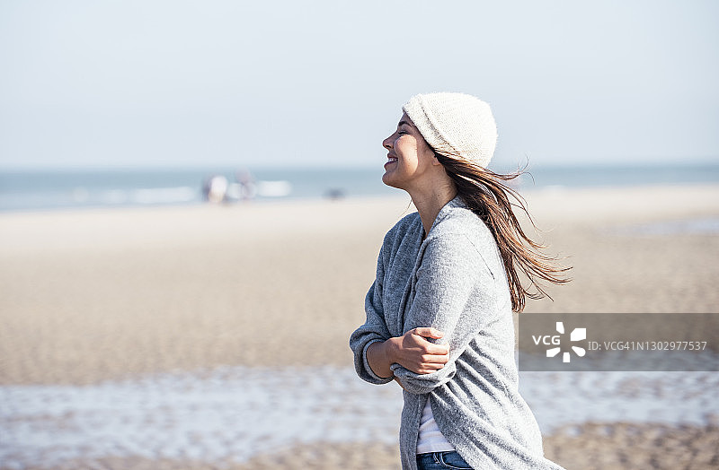 戴针织帽的快乐年轻女人在海滩拥抱自己图片素材