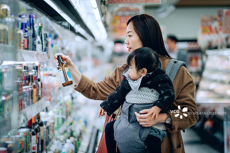 年轻的亚洲母亲和可爱的小女儿一起购买杂货。她正在看超市冷藏通道里的一瓶饮料图片素材