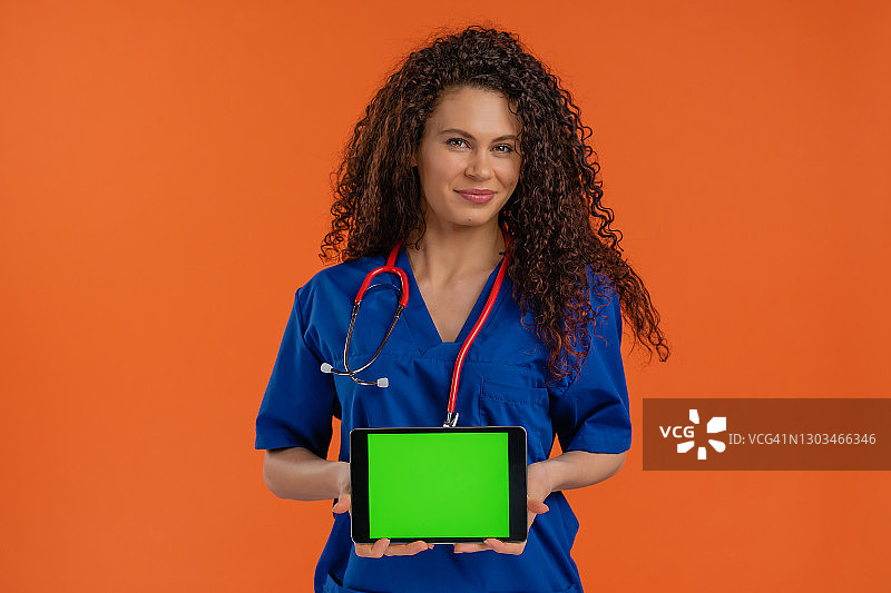 快乐的女医生展示带有色度键绿屏的数字平板电脑，情绪积极，橙色背景图片素材