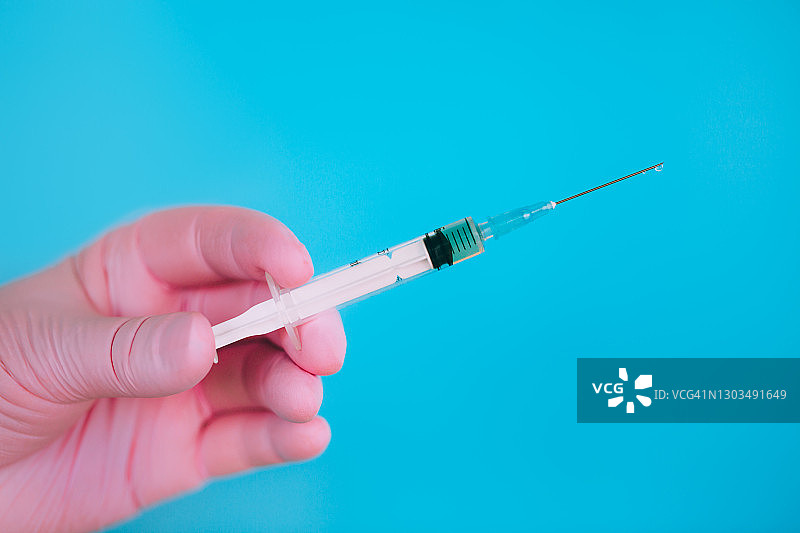 戴粉色手套的医生的手拿着小注射器，蓝色背景上有冠状病毒疫苗图片素材