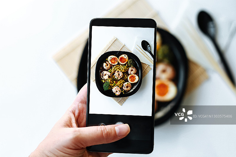 拍摄日式亚洲传统食物的电话屏幕特写：拉面，含煮鸡蛋、面条、虾和西兰花。图片素材