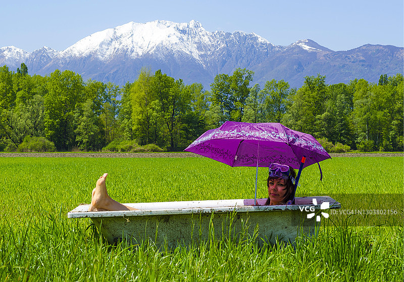 女人躺在浴缸里，戴着雨伞和潜水面罩，背景是绿色田野、雪山和树木图片素材