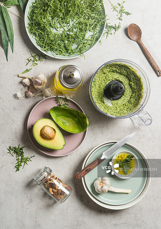 灰色厨房桌上自制绿色鹰嘴豆泥，配牛油果、芝麻菜、大蒜、橄榄油和坚果等食材。俯视图图片素材