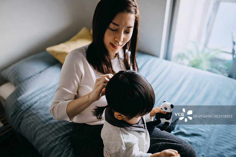 出门前，亚洲母亲在床上为小女儿扎头发并穿好衣服，家庭生活方式图片素材