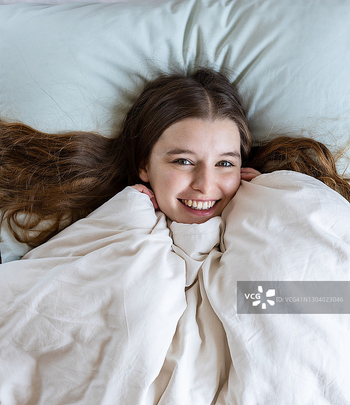 清晨，害羞的金发年轻女人躲在被窝里对着镜头微笑图片素材
