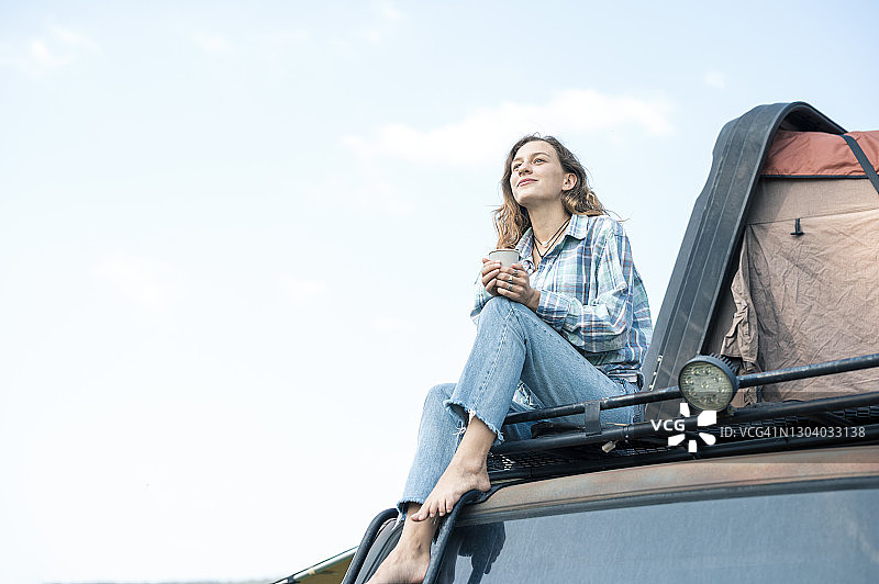 坐在露营车顶上手持咖啡杯欣赏风景的快乐女子肖像——可持续生活方式图片素材