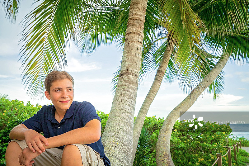 坐在高高的棕榈树干上，眺望大海的青少年男孩图片素材