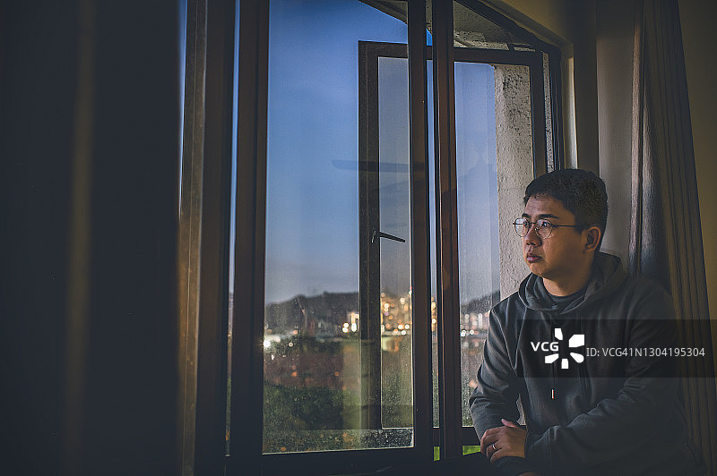 华人中年男子日落时在家中望着窗外，表情严肃，穿着连帽衫图片素材