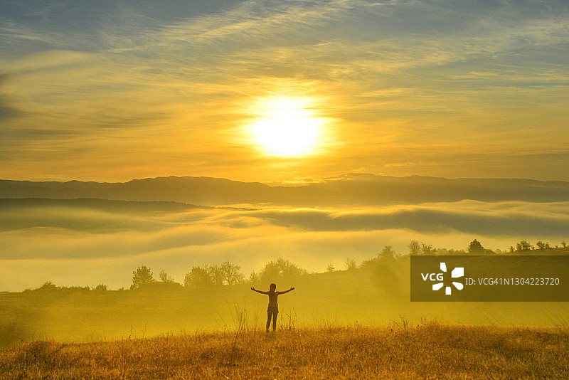 在云雾缭绕山谷中日出时分张开双臂的女子剪影图片素材