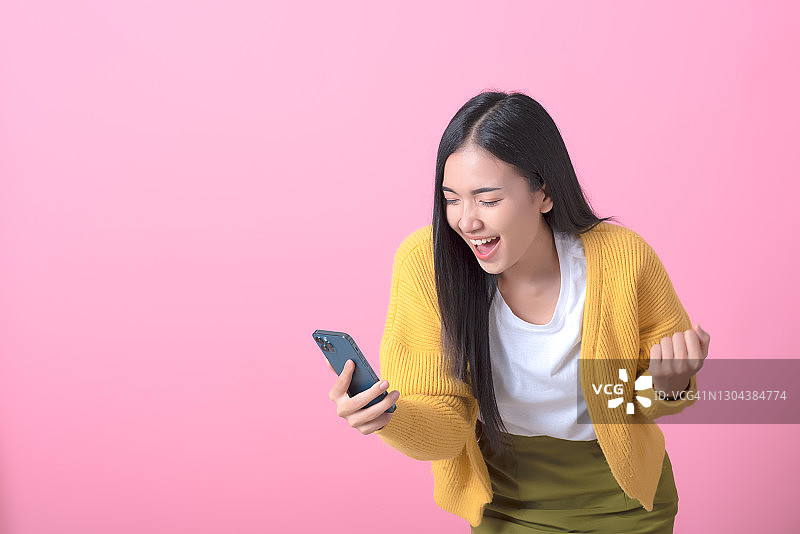 快乐的亚洲年轻女人使用智能手机，收到手机聊天应用上的好消息，粉色背景图片素材
