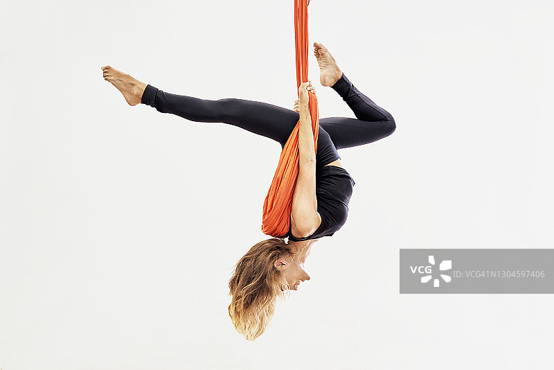金发女子悬挂在橙色秋千上练习空中瑜伽的正面图图片素材