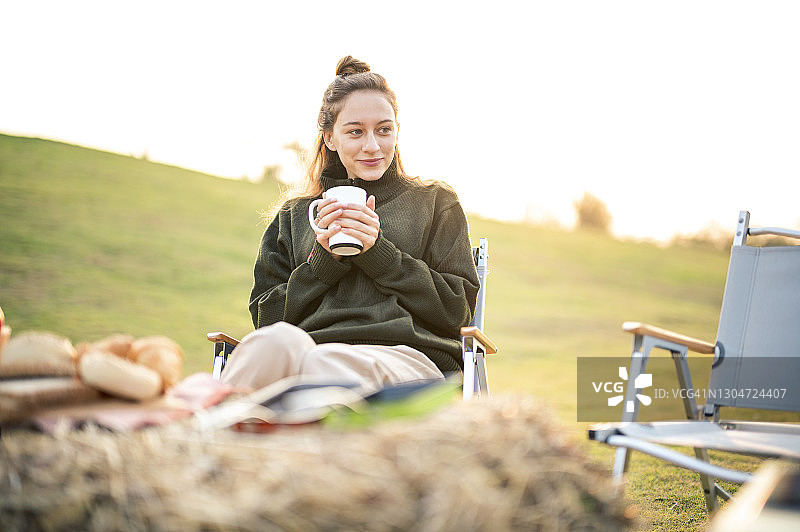 女性在日出时坐在露营椅上享用咖啡或热饮。露营，简单，可持续的生活方式。图片素材