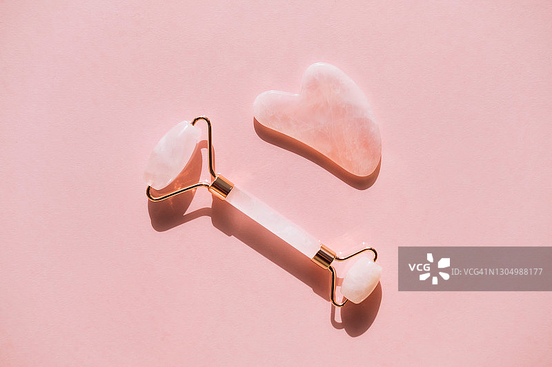 粉色背景上的粉色石英滚轮和刮痧板，用于面部按摩。年度潮流产品，健康保健概念图片素材