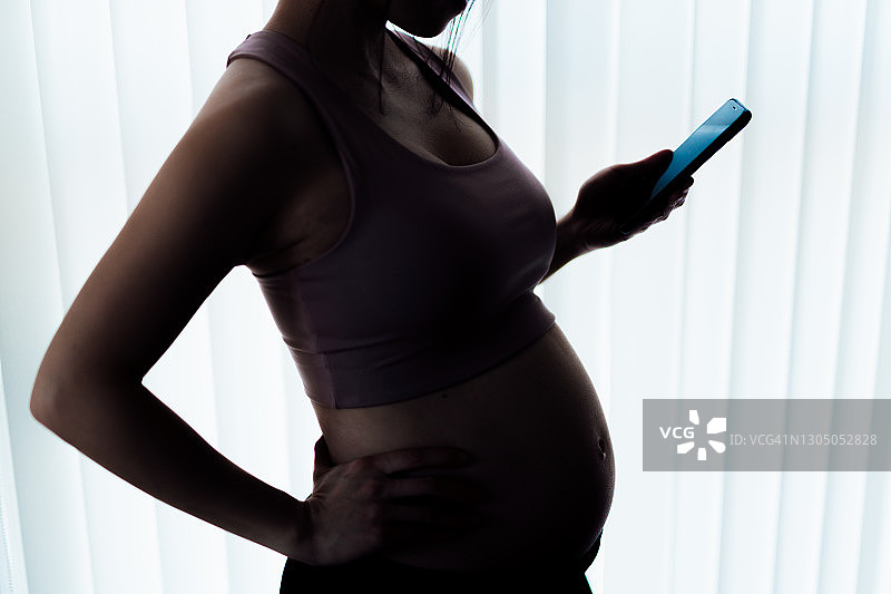 孕妇手持手机站在家中窗边的侧面轮廓图图片素材