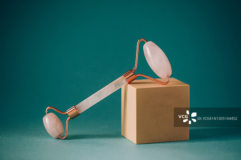 在绿松石背景的米色立方体形状领奖台上的面部按摩用粉色石英滚轮。健康概念图片素材