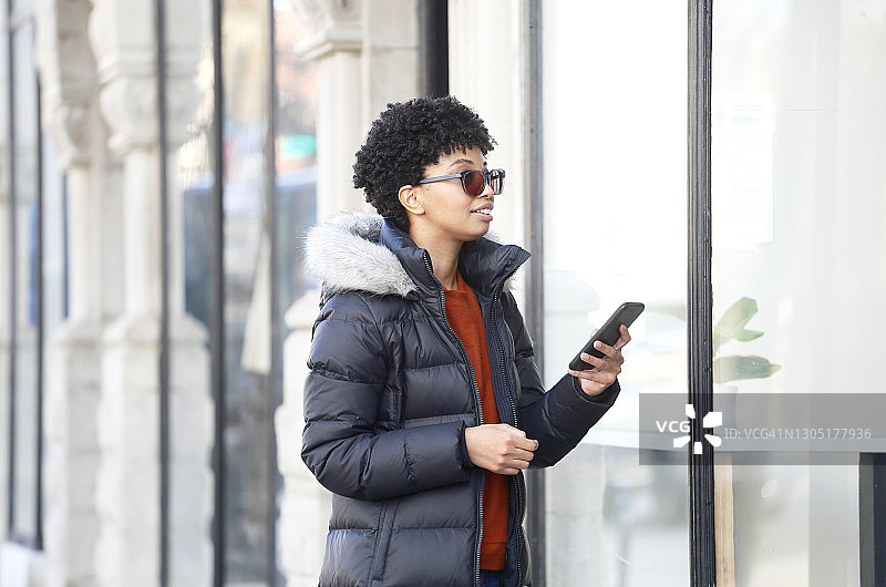 一位戴着眼镜、身穿休闲冬装的非裔美国年轻女性站在商店前使用手机图片素材