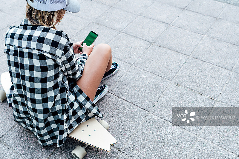 穿着短裤和帽子，坐在长板滑板上的金发女人看着带色度键屏幕的手机图片素材
