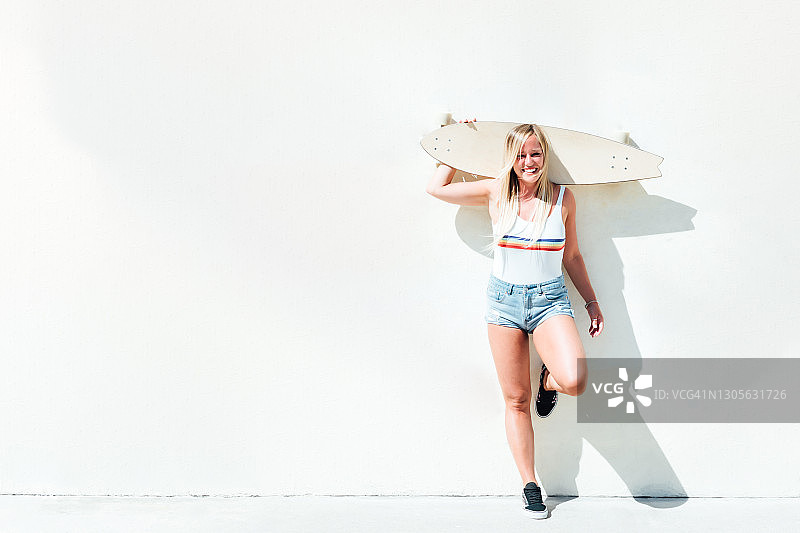身穿短裤和条纹衬衫的金发女人肩扛长板滑板倚墙而立图片素材