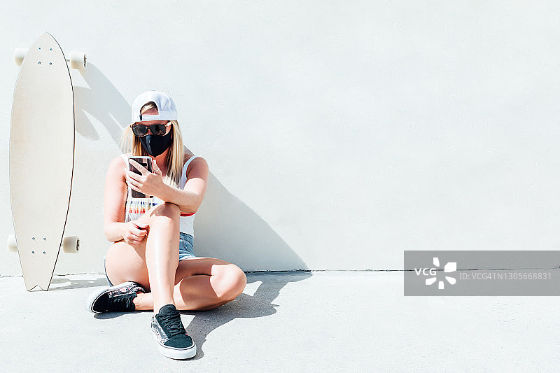 金发女人戴着口罩、黑色太阳镜和灰色帽子，坐在地板上看手机，长板滑板靠在墙上图片素材