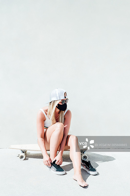 戴着口罩、太阳镜和灰色帽子，坐在长滑板上的金发女人图片素材