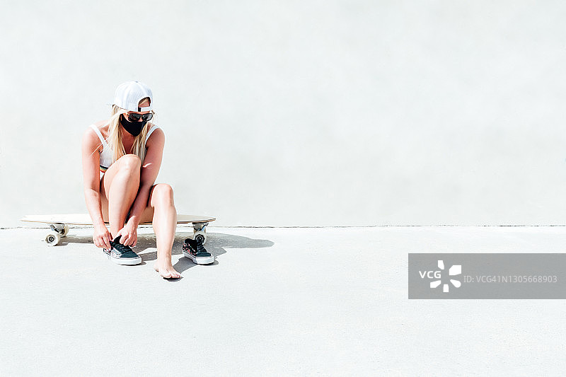 戴着口罩、太阳镜和灰色帽子，坐在长滑板上的金发女人图片素材