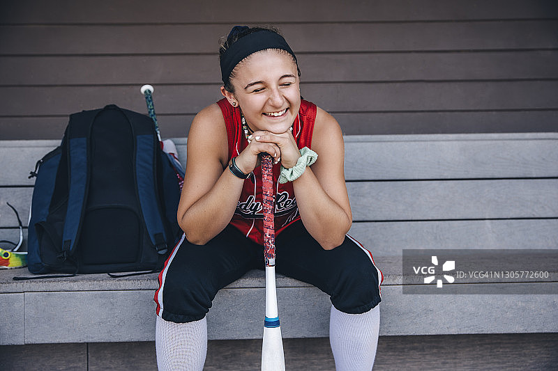 靠在球棒上坐在休息区里的快乐女子垒球运动员图片素材