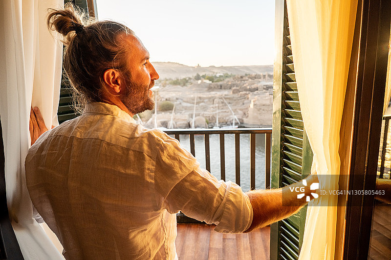 日出时，男子在酒店房间里拉开窗帘，尼罗河图片素材