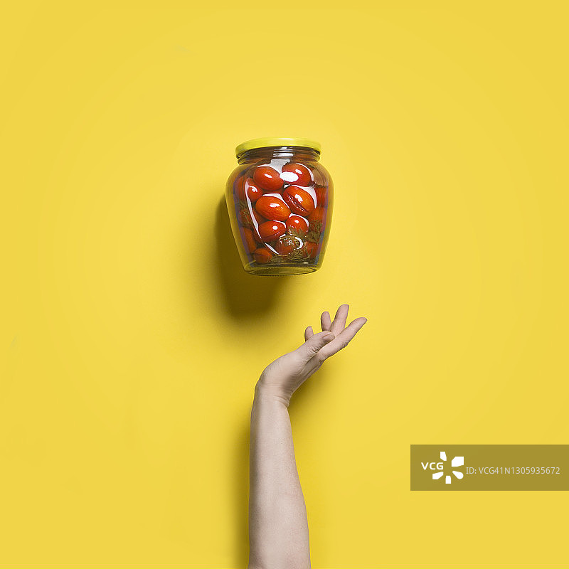 黄色背景下，飞舞的玻璃罐头瓶装着腌制红番茄和女人手图片素材