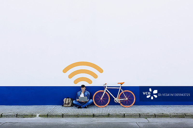 休闲装男子坐在人行道上，在白色墙壁的Wi-Fi标志旁用笔记本电脑图片素材