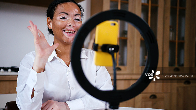 年轻拉丁裔白癜风女性在厨房餐桌旁用智能手机进行视频通话图片素材