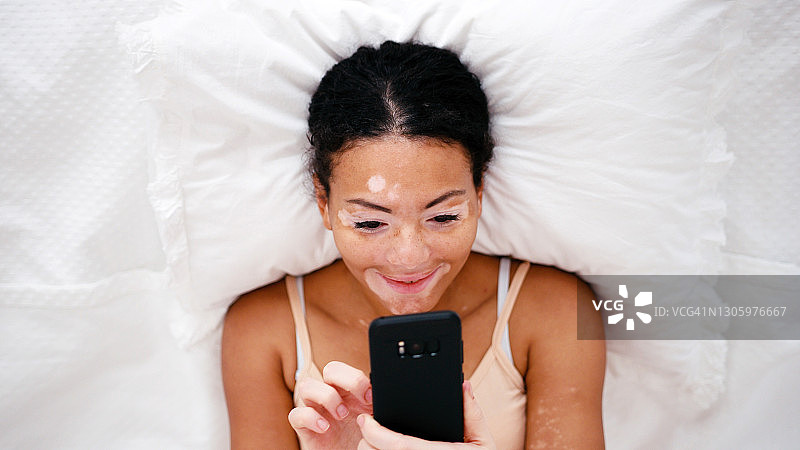 年轻拉丁裔白癜风女性在床上滑动手机图片素材