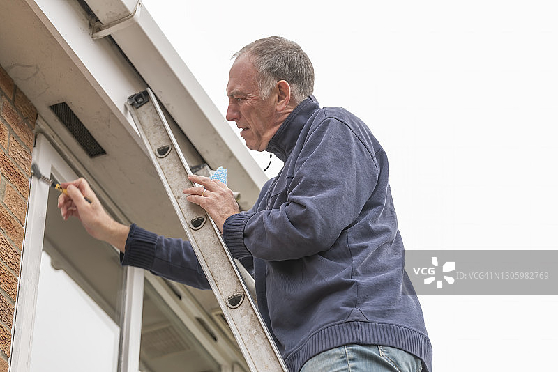 年长的男人清洁并在泄漏的窗户周围涂抹密封剂图片素材