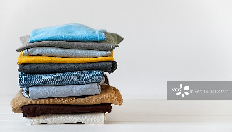 白色桌子或背景上的一堆物品：干净的折叠棉质牛仔服装和色彩鲜艳的T恤。家务、熨烫、护理和物品存储的概念。文案空间。图片素材